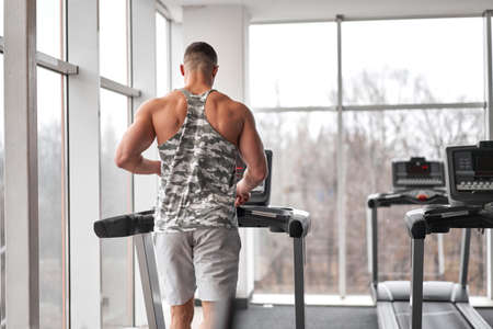 Muscular athletic bodybuilder fitness model running treadmill gym near big window jogging stationary...