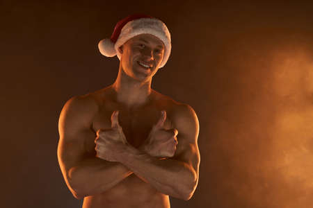 Portrait of muscular man wearing Christmas Santa hat, folded hands and smile on smoky background Macho Shirtless Naked torso stripper Hot sexy Santa guy Seductive male for holidayの写真素材