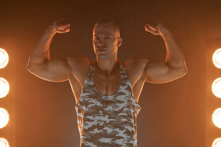 Active lifestyle concept. Professional bodybuilder showing biceps and smiling at camera, lamps...
