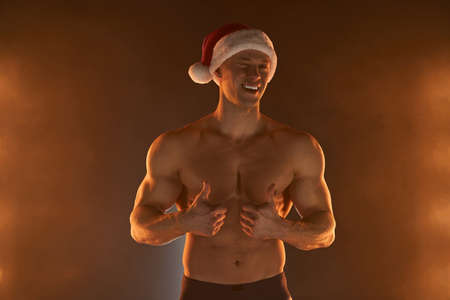 Portrait of muscular man wearing Christmas Santa hat, showing thumb up and smile on smoky background Macho Shirtless Naked torso stripper Hot sexy Santa guy Seductive male for holidayの写真素材