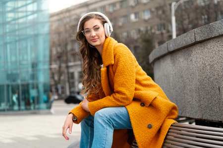 Woman Eyeglasses Listen Music Headphone Outdoor Sitting Bench Outdoor Dressed Stylish Yellow Coat Smile Caucasian Female 30s Enjoy Podcast Or Audio Books Outsideの写真素材