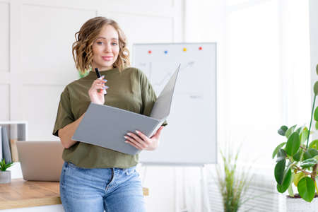 Businesswoman holding folder for paper write marker thinking dreaming standing near office table. Business person femal ypoung adult caucasian on modern white interiorの写真素材