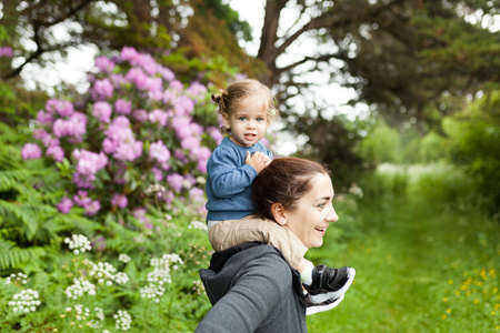 Little caucasian girl sitting shoulders his mother. Family with one parent walking nature flower...