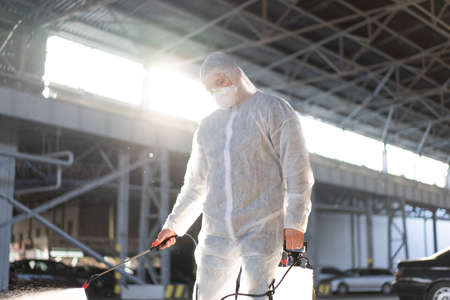 Man dressed white protective overalls spraying surface antibacterial sanitizer sprayer during quarantine Caucasian person protective suit disinfects parking during  pandemic backlightの写真素材