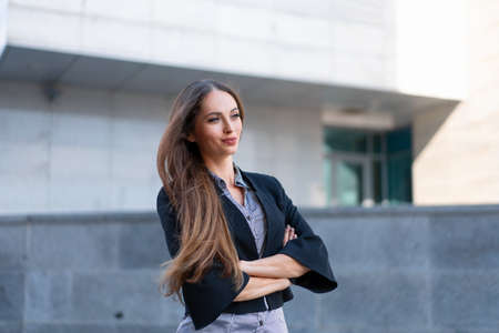 Business woman with long hair dressed black jacket standing outdoor near corporate office building hands folded Beautiful caucasian female business person portrait Side view looking awayの写真素材