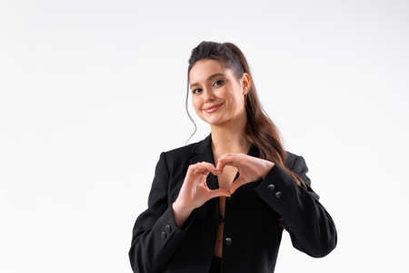 Young business woman showing heart gesture by hand dressed black jacket standing studio isolated on white background. Caucasian female business person looking at camera and smile. Medium shootの写真素材