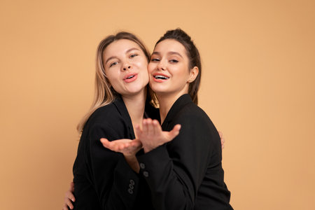 Two young business woman dressed black suit embrace laugh and happy standing in studio isolated over peach color background. Female friends and partners in business concept sending blow kiss in cameraの写真素材