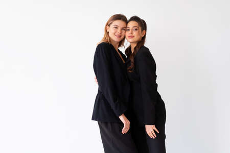 Concept of partnership in business. Two Young businesswoman dressed black formal suit standing studio isolated white background. Caucasian female business person cheek to cheek smileの写真素材