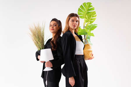 Two business woman dressed black suit holding houseplant and lamp standing isolated on white...