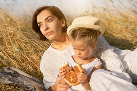 Mother and daughter sitting picnic high yellow grass child eat bun. Happy family with one child...