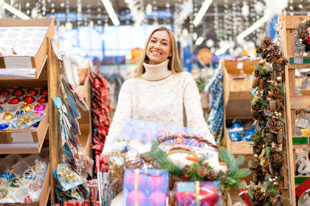 Christmas decoration shopping. Caucasian woman choose new year decor in shopping mall. Medium age female dressed white sweater buying christmas ornament toys indoor storeの写真素材