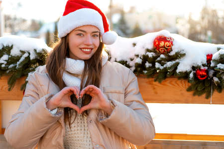 Winter portrait young adult beautiful woman in Santa hat showing heart shape by fingers. Christmas...