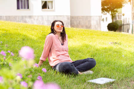 Photo of european woman 25s sitting on green grass in park with legs crossed during summer day while using laptop. Caucasian female hipster with dreadlocks and pink sunglasses on use laptop freelancerの写真素材