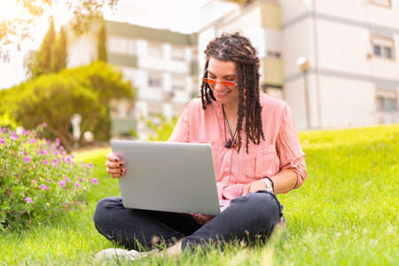 Photo of european woman 25s sitting on green grass in park with legs crossed during summer day while using laptop. Caucasian female hipster with dreadlocks and pink sunglasses on use laptop freelancerの写真素材