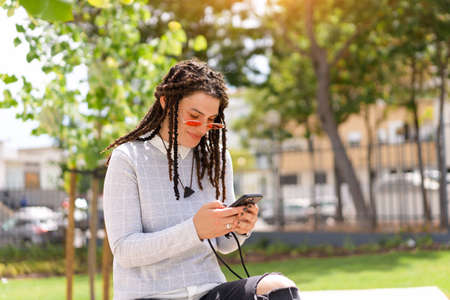 Young woman hipster with dreadlocks in pink sunglasses use smartphone sitting summer park outdoor....