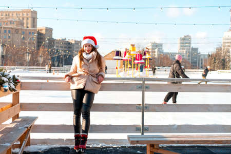 Beautiful lovely young adult woman brunet hair warm winter jackets stands near ice skate rink background Town Square. Christmas mood lifestyle Happy holiday woman walk snowy day Winter leisureの写真素材