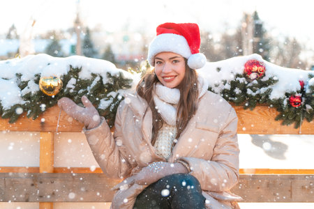 Winter portrait young adult beautiful woman in Santa hat. Christmas mood. snowing. Winter beauty fashion concept. Pretty young woman sitting outdoor near Christmas garlandの写真素材