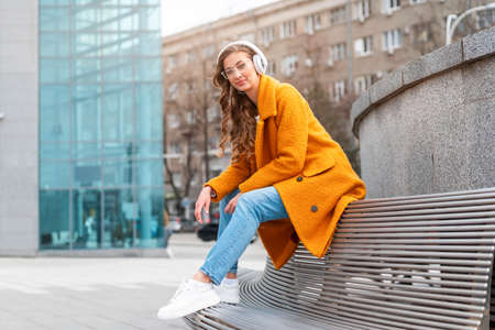 Woman Eyeglasses Listen Music Headphone Outdoor Sitting Bench Outdoor Dressed Stylish Yellow Coat Smile Caucasian Female 30s Enjoy Podcast Or Audio Books Outsideの写真素材