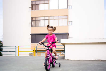 Cute little smiling girl riding bike bicycle in city on parking sunny summer day. Active family leisure with kids. Little caucasian girl 5 years old have fun with pink child bicycleの写真素材