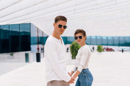 Smiling beautiful woman and her handsome boyfriend. Woman in casual summer jeans. Happy cheerful couple in sunglasses holding hand each other. Couple posing on the street modern building backgroundの写真素材