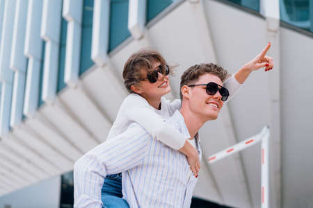 Carefree young urban couple in sunglasses doing piggyback at city street outdoor with modern commercial building on background. Man carrying young woman on his back, woman show by finger awayの写真素材
