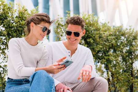 Modern Businessman showing something in the smartphone to his female assistant. Modern business young business people in sunglasses using smartphone smile sitting bench outdoor in office districtの写真素材