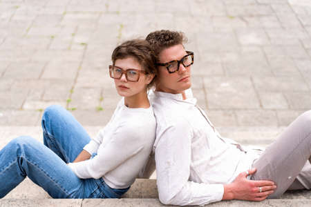 Portrait of two attractive students man and woman wearing stylish glasses sitting floor outdoor and...