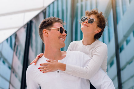Carefree young urban couple in sunglasses doing piggyback at city street outdoor with modern commercial building on background. Man carrying young woman on his back, positive emotionの写真素材