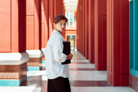 businesswoman portrait. Caucasian female business person walking outdoor with business paper document holder. Young beautiful modern business woman back view. Confidence employer. looking at cameraの写真素材