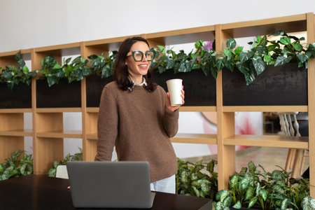 Happy European Female Entrepreneur Standing Near Desk With Laptop Computer Posing Smiling In Modern Office Indoor. Freelance Career And Successful Entrepreneurship Conceptの写真素材