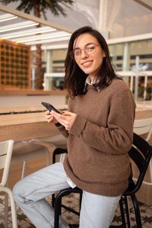 Portrait of positive cheerful girl use smart phone look at camera share social media news wear stylish trendy jumper sitting bar stool. female smartphone user. woman in glasses holding smartphoneの写真素材