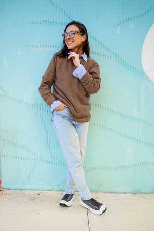 Happy woman in glasses outdoor on blue color background. positive people concept. Smiling girl looking away, hands in pocket, dressed sweater and jeans. vertical full lengthの写真素材
