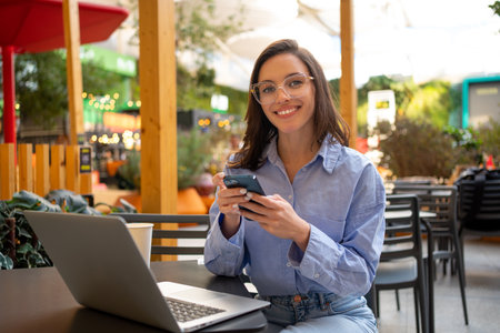 Freelance worker doing remote work using smartphone internet. looking at camera and smile. Business woman with mobile phone in outdoor cafeteria, restaurant on social media app online.の写真素材
