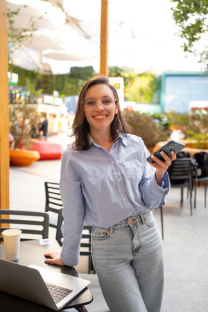 Freelance worker doing remote work using smartphone and laptop, looking at camera and smile. Female freelancer with mobile phone standing in outdoor cafe holding phone in hand. vertical shootの写真素材