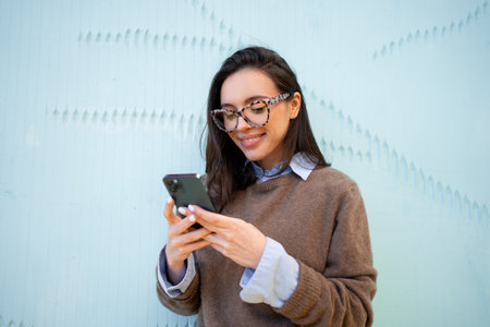 Satisfied hipster girl wear glasses, types text message on mobile phone, enjoys online communication, types feedback, wears sweater, isolated on blue color background. technology conceptの写真素材