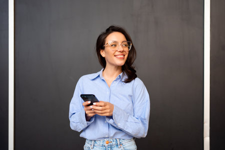 Portrait smiling woman holding smartphone in hand looking away, wear glasses, types text message on mobile phone, enjoys online communication, on black studio background. communication conceptの写真素材