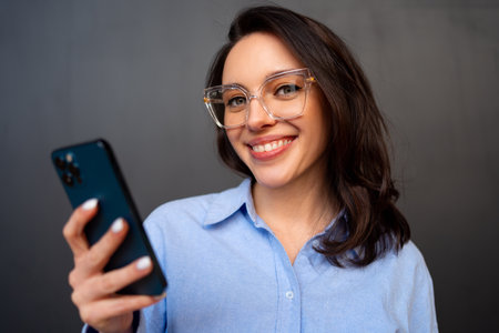 Portrait smiling woman holding smartphone in hand looking at camera, wear glasses, types text message on mobile phone, enjoys online communication, on black studio background.の写真素材