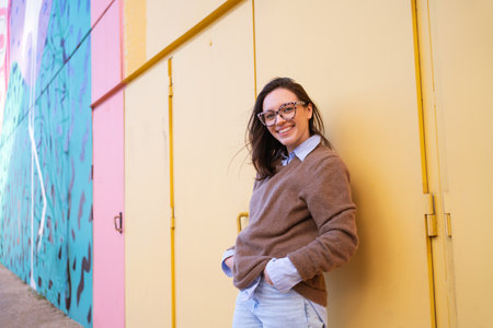 Happy woman in glasses outdoor on yellow color background side view. positive people concept. Smiling girl looking at camera, hands in pocket, dressed sweater and jeansの写真素材