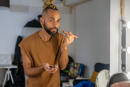 Handsome bearded black gay applying make up on face looking mirror. Transgender non binary person use makeup brush.の写真素材