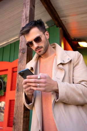Male adult wearing sunglasses using smartphone for sending message. Man in stylish trench coat and fashionable eyewear texting on cellphone. Modern lifestyle, connection conceptの写真素材