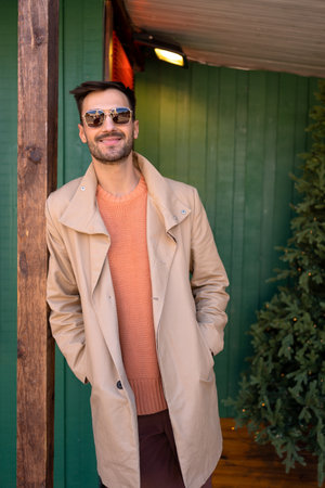 Handsome man in beige stylish trench in front of wooden house. Elegant male wearing salmon-colored sweater and pale brown trench coat standing outdoors. Fashionable guy in sunglasses winter holidaysの写真素材