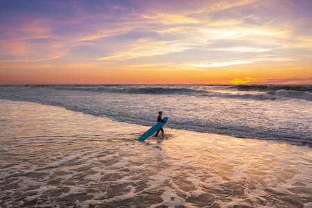 Surfer walking in ocean on sunset with surfboard. Person with surfboard stands in ocean water. Landscape with pink and violet clouds, ocean waves and sand beach. Surfer wathing the wavesの写真素材