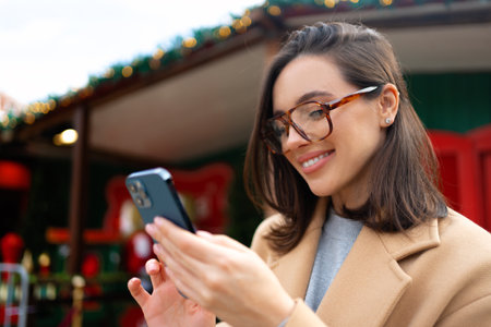 Woman in glasses using mobile phone at day in the city. Happy woman using smartphone dressed stylish trench coat looking device screenの写真素材