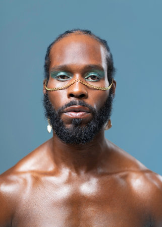 Black gay stands isolated on blue background and stares into camera with serious expression on face. Close-up portrait of handsome adult transgender with blue eyeshadows and golden accessory on faceの写真素材