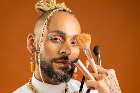 Latino gay make up artist holding makeup brush. Handsome guy wear bright make-up on isolated orange background. Fashion lgbt concept. Handsome makeup artist close up portraitの写真素材