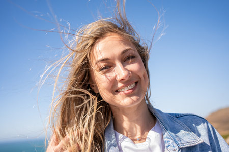 Closeup portrait of happy smiling beautiful woman with blond windswept hair. Cheerful female tourist on sea shore against blue sky. Lady enjoys summer vacation.の写真素材