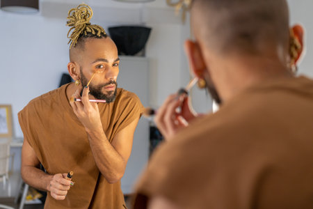 Black male gay applying make up looking mirror standing back to camera. Stylish homosexual man in dressing room doing make-up on faceの写真素材