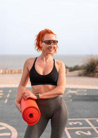Smiling redhead woman in sunglasses holding rolled yoga mat and looking away. Fit sporty female athlete in activewear looking away at summer park. Fitness and health concept.の写真素材