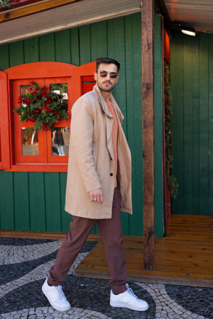Full body of confident handsome man in sunglasses and trendy beige trench coat walking outside Christmas decorated house. Side view of hipster male in fashionable outfit in city.の写真素材