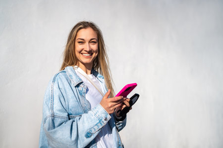 Portrait of cheerful young woman in denim shirt holding smartphone and looking at camera. Smiling female tourist standing against white wall on sunny day.の写真素材